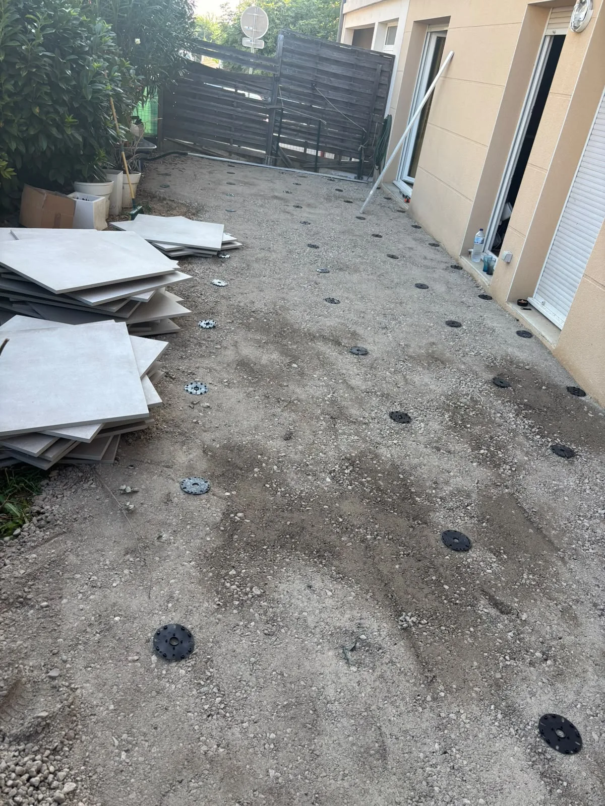 Avant — sol en gravier avec les plots réglables répartis avant la pose, et les dalles de grès cérame empilées sur le côté